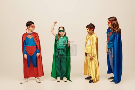 happy interracial kids in colorful costumes looking at girl in green superhero outfit standing with raised hand and protesting on grey background in studio, Child Protection Day concept 