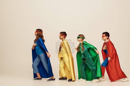 side view of happy interracial kids in colorful costumes with cloaks and masks smiling and walking together on grey background in studio, Child Protection Day concept  mug #655802130