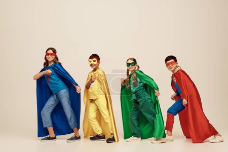 happy interracial kids in colorful costumes with cloaks and masks smiling and walking together while looking at camera on grey background in studio, Child Protection Day concept 