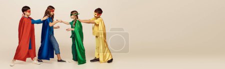 Téléchargez les photos : Vue de côté des enfants multiculturels en costumes de super-héros colorés avec des masques et des manteaux se battant les uns avec les autres sur fond gris en studio, concept de journée internationale de l'enfant, bannière - en image libre de droit