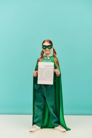 glückliches Mädchen in grünem Superheldenkostüm mit Mantel und Maske, das mit leerem Papier steht und in die Kamera schaut, während es den Kinderschutztag auf blauem Hintergrund feiert 