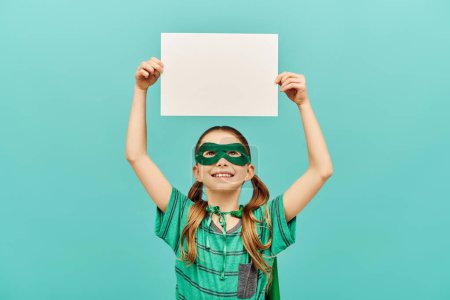 happy preteen girl in green superhero mask holding blank paper above head and looking up on blue background, World child protection day concept 