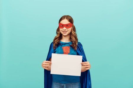 niña preadolescente positiva en traje de superhéroe con máscara roja de pie con papel en blanco y mirando a la cámara en el fondo azul, concepto del día mundial de la protección del niño 