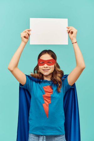 Unbekümmertes Mädchen im Superheldenkostüm mit Mantel und roter Maske, leeres Papier über dem Kopf und Blick in die Kamera auf blauem Hintergrund, Happy Children 's Day Konzept 