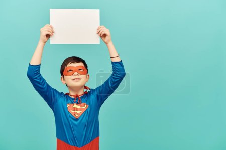 Frühchen asiatischer Junge im Superheldenkostüm mit Maske, leeres Papier über dem Kopf und Blick nach oben auf blauem Hintergrund, Konzept zum Internationalen Kinderschutztag 
