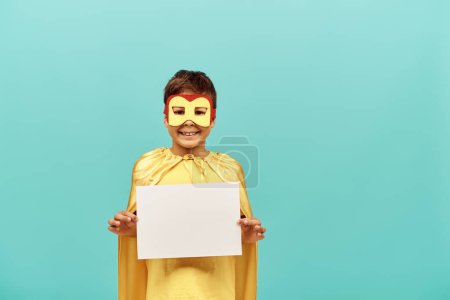 niño multirracial sonriente en traje de superhéroe amarillo con máscara sosteniendo papel en blanco sobre fondo azul, concepto del día de los niños felices 
