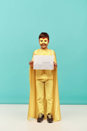 in voller Länge positiver multirassischer Junge im gelben Superheldenkostüm mit Maske, die leeres Papier auf blauem Hintergrund hält, Konzept zum Internationalen Kinderschutztag 