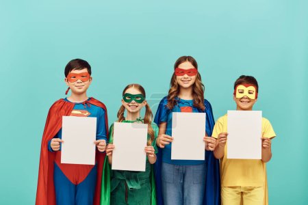 happy interracial kids in colorful superhero costumes with masks smiling and holding blank papers while looking at camera on blue background in studio, Child Protection Day concept 