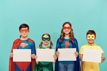 lächelnde Kinder in farbenfrohen Superheldenkostümen mit Masken, die leere Papiere halten, während sie in die Kamera auf blauem Hintergrund im Studio schauen, Happy Children 's Day Konzept