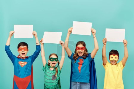 szczęśliwy międzyrasowe dzieci w kolorowe kostiumy superbohatera z maskami trzymając puste papiery nad głowami patrząc na aparat na niebieskim tle w studio, koncepcja Światowego Dnia Ochrony Dzieci 