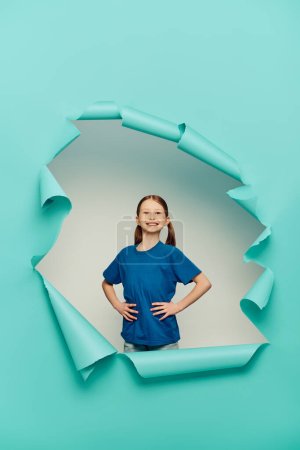 glückliches rothaariges Mädchen im T-Shirt, das in die Kamera blickt, während es mit den Händen auf den Hüften um ein blaues zerrissenes Papierloch auf weißem Hintergrund posiert, Konzept zum Internationalen Kinderschutztag 