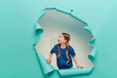 shocked redhead preteen girl in t-shirt looking at blue torn paper creating hole on white background, International Child Protection Day concept 
