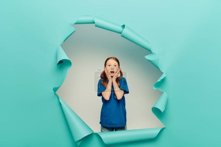 scared redhead preteen girl in t-shirt looking at camera while touching face with hands around blue torn paper hole on white background, Global Children Day concept 