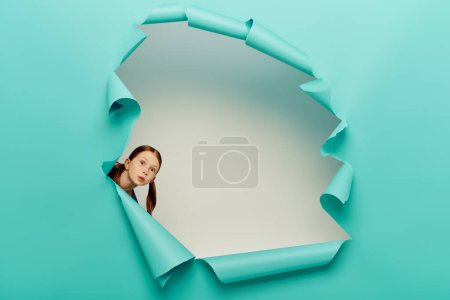 Foto de Chica preadolescente pelirroja sorprendida mirando a la cámara a través de agujero de papel rasgado azul sobre fondo blanco, concepto del Día Internacional de la Protección del Niño - Imagen libre de derechos
