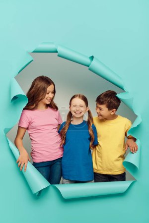 Lächelnde Kinder in bunten T-Shirts schauen beim Kinderschutztag ihren rothaarigen Freund an und stehen hinter einem Loch im blauen Papierhintergrund