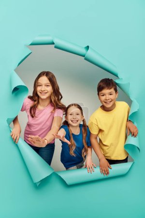 Positive multiethnic kids in colorful t-shirts outstretching hands at camera while celebrating international children day near hole in blue paper background