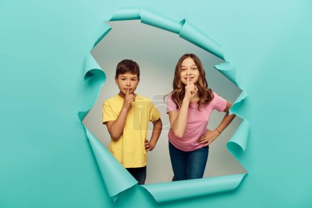 Smiling girl holding hand on hip and showing secret gesture near multiracial friend while celebrating child protection day behind hole in blue paper background