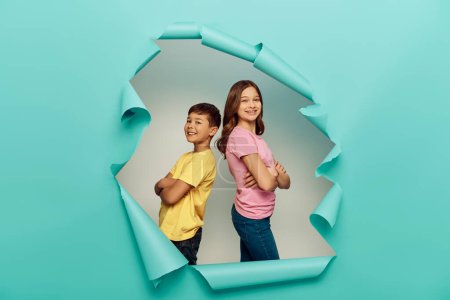 Smiling interracial preteen kids in colorful t-shirts crossing arms while standing back to back and celebrating child protection day behind hole in blue paper background