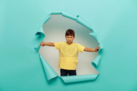 Angry multiracial boy in yellow t-shirt looking at camera during international child protection day while standing behind hole in blue paper on white background