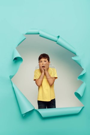 Shocked multiracial boy in yellow t-shirt touching cheeks and looking at camera during world child protection day while standing behind hole in blue paper background