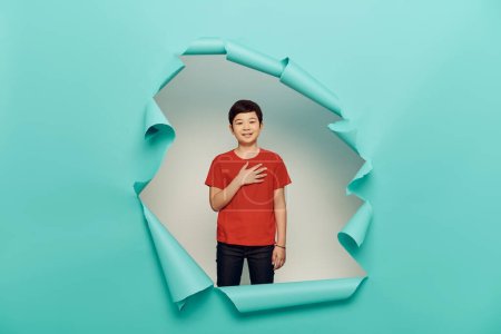 Sonriente asiático preadolescente chico en rojo camiseta mirando a la cámara durante la celebración del día de la protección del niño y de pie detrás del agujero en fondo de papel azul