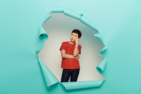 Präventiver asiatischer Bub im roten T-Shirt, der wegschaut, während er hinter einem Loch in blauem Papier auf weißem Hintergrund steht, Konzept zum Weltkinderschutztag 