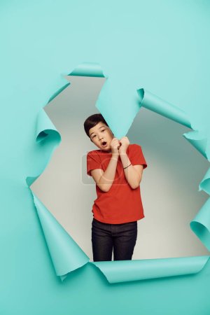 Shocked asiático preadolescente chico rojo camiseta mirando cámara y tocando agujero en azul papel mientras celebrando el día de la protección del niño en blanco fondo