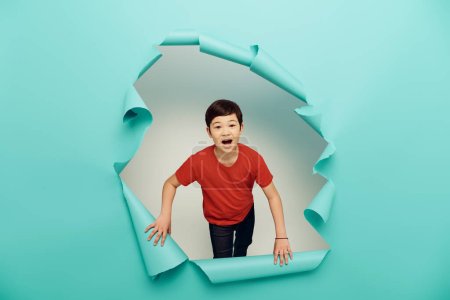 Astonished asian preteen kid in casual red t-shirt looking at camera during child protection day celebration behind hole in blue paper on white background