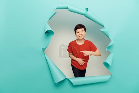positiver asiatischer Bub in rotem T-Shirt zeigt mit dem Finger während Kinderschutztag Feier hinter Loch in blauem Papier und auf weißem Hintergrund