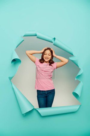 Positive preteen girl in pink t-shirt touching hair while standing behind hole in blue paper on white background, Happy children's day concept 