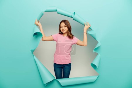 Carefree preteen girl in casual clothes looking at camera during child protection day celebration behind hole in blue paper on white background