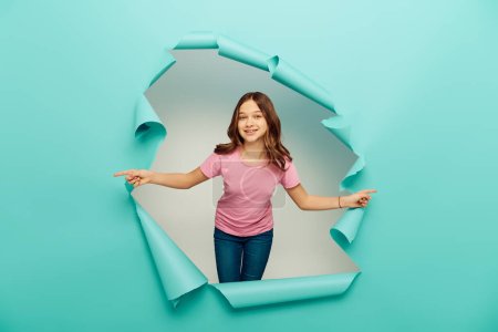 Joyful preteen girl in casual clothes pointing with fingers during international children day celebration behind hole in blue paper background
