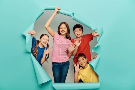 Lächelnde Kinder in lässiger Kleidung winken während der internationalen Kindertagsfeier hinter einem Loch im blauen Papierhintergrund in die Kamera