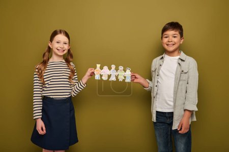 Smiling multiethnic preteen friends in casual clothes holding paper drawn characters and looking at camera during child protection day celebration on khaki background