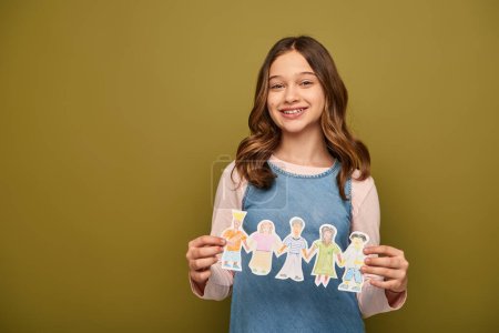 Smiling preteen girl in denim sundress holding drawn paper characters and looking at camera during child protection day celebration on khaki background