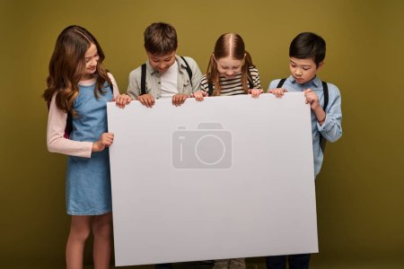 positive multiethnic preteen kids with backpacks smiling while standing together and  looking at empty placard on khaki background, Happy children's day concept 