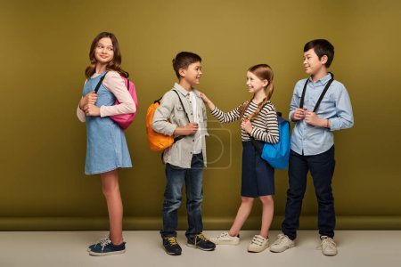 Pleine longueur d'enfants interracial souriants avec des sacs à dos regardant des amis parler pendant la célébration de la journée internationale de la protection de l'enfance sur fond kaki