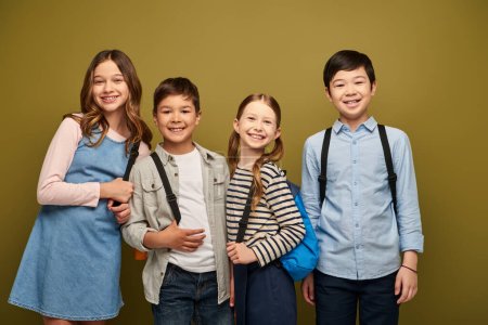 Lächelnde multiethnische Kinder mit Rucksäcken in lässiger Kleidung, die in die Kamera schauen, während sie bei der Feier zum Kinderschutztag auf khakifarbenem Hintergrund zusammenstehen