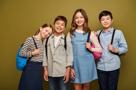 Positive interrassische und frühkindliche Kinder in Freizeitkleidung mit Rucksäcken und Blick in die Kamera bei der Kinderschutztagsfeier auf khakifarbenem Hintergrund