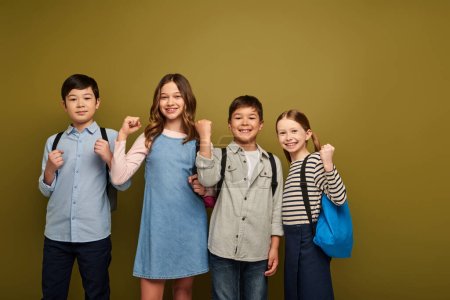 Smiling multiethnic preteen kids with backpacks showing fists as power gesture and looking at camera during world child protection day celebration while standing together on khaki background