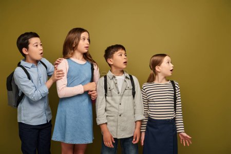 Schockierte multiethnische Vorschulkinder in lässiger Kleidung mit Rucksäcken, die wegschauen und den Mund aufmachen während der Kinderschutztagsfeier auf khakifarbenem Hintergrund