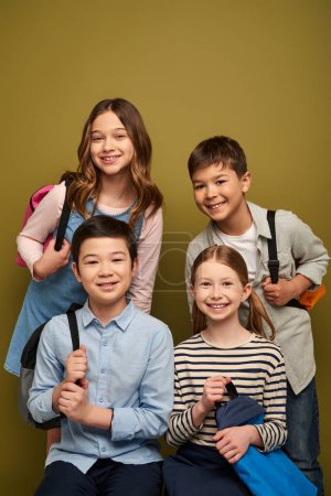 Lächelnde multiethnische Kinder in lässiger Kleidung mit Rucksäcken und Blick in die Kamera während der Kinderschutztagsfeier auf khakifarbenem Hintergrund