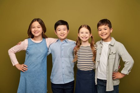 Lächelnde multiethnische Kinder umarmen Freunde in lässiger Kleidung, blicken gemeinsam in die Kamera und posieren während der Feier zum Kinderschutztag auf khakifarbenem Hintergrund