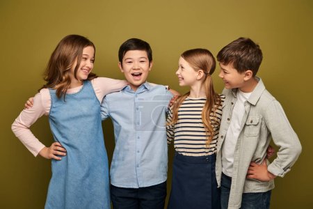 Happy interracial kids in casual clothes hugging and looking at asian friend with opened mouth while celebrating international child protection day on khaki background