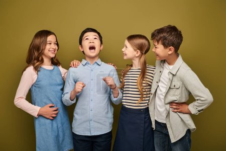 Excited asian preteen boy screaming and standing near happy multiethnic friends in casual clothes during child protection day celebration on khaki background