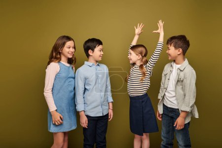 Smiling redhead girl in casual clothes standing with raised hands, gesturing and scaring cheerful multiethnic friends while celebrating child protection day on khaki background