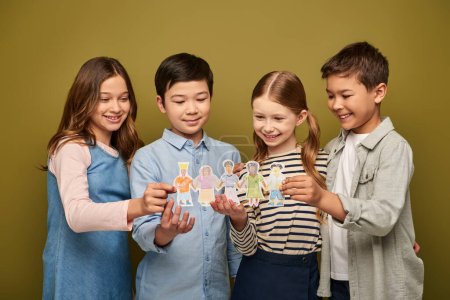 Smiling interracial preteen kids in casual clothes with friends holding drawn paper characters while celebrating child protection day on khaki background
