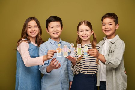 Preteen kids in legerer Kleidung halten gezeichnete Papierfiguren in der Hand und lächeln in die Kamera während der Feier zum internationalen Kinderschutztag auf khakifarbenem Hintergrund