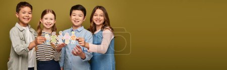 Happy interracial preteen friends in casual clothes holding drawn paper craft characters and looking at camera during child protection day celebration on khaki background with copy space, banner 