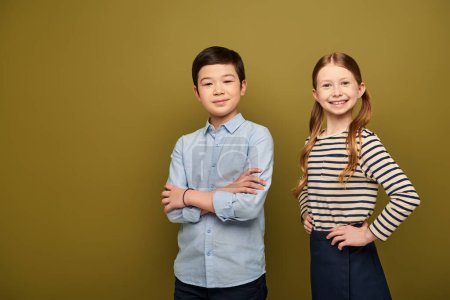 Smiling redhead girl holding hands on hips and looking at camera near asian friend crossing arms during child protection day celebration on khaki background